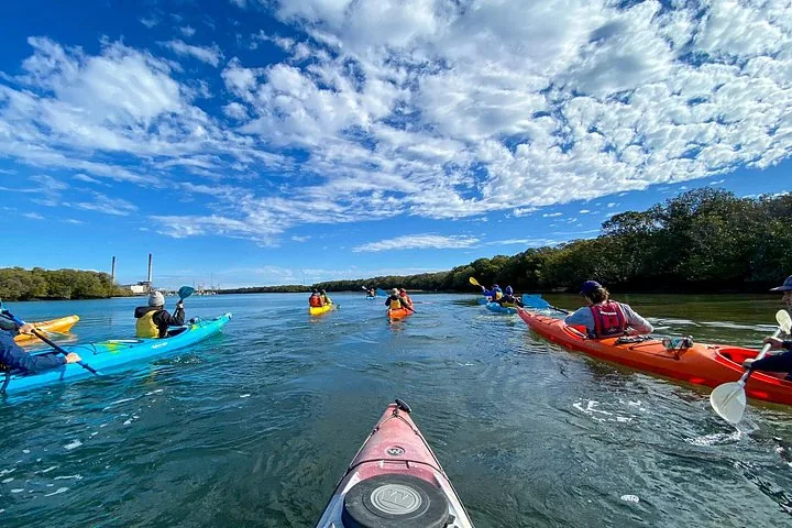 Kayakers following a leader on a calm river with industrial buildings in the distance