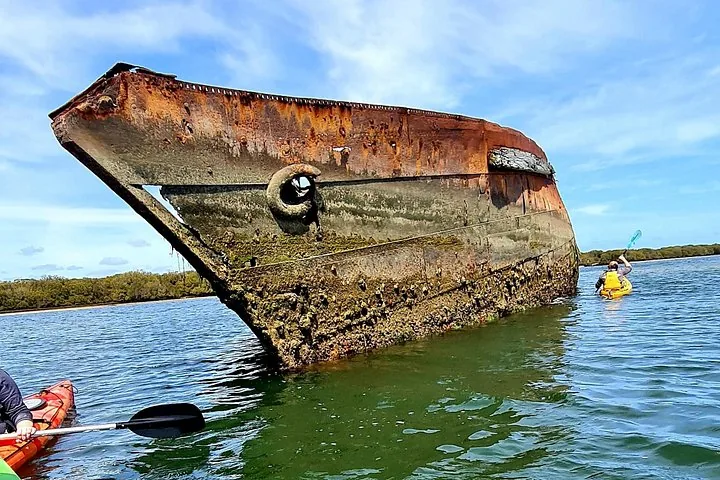 Un kayakiste pagayant devant une grande épave abandonnée et rouillée dans l'eau